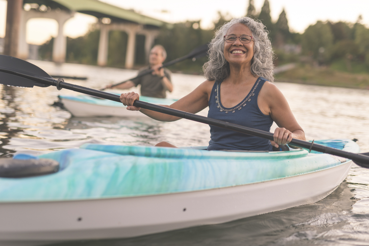 A mixed race senior couple is kayaking down a river together. The image's focus is on the elderly woman smiling in the foreground. She is Pacific Islander and has graying hair. Her husband is seen in a kayak in the background. In the distance behind the couple you can see the shoreline and a bridge. The happy couple are wearing casual clothing. It is warm outside.